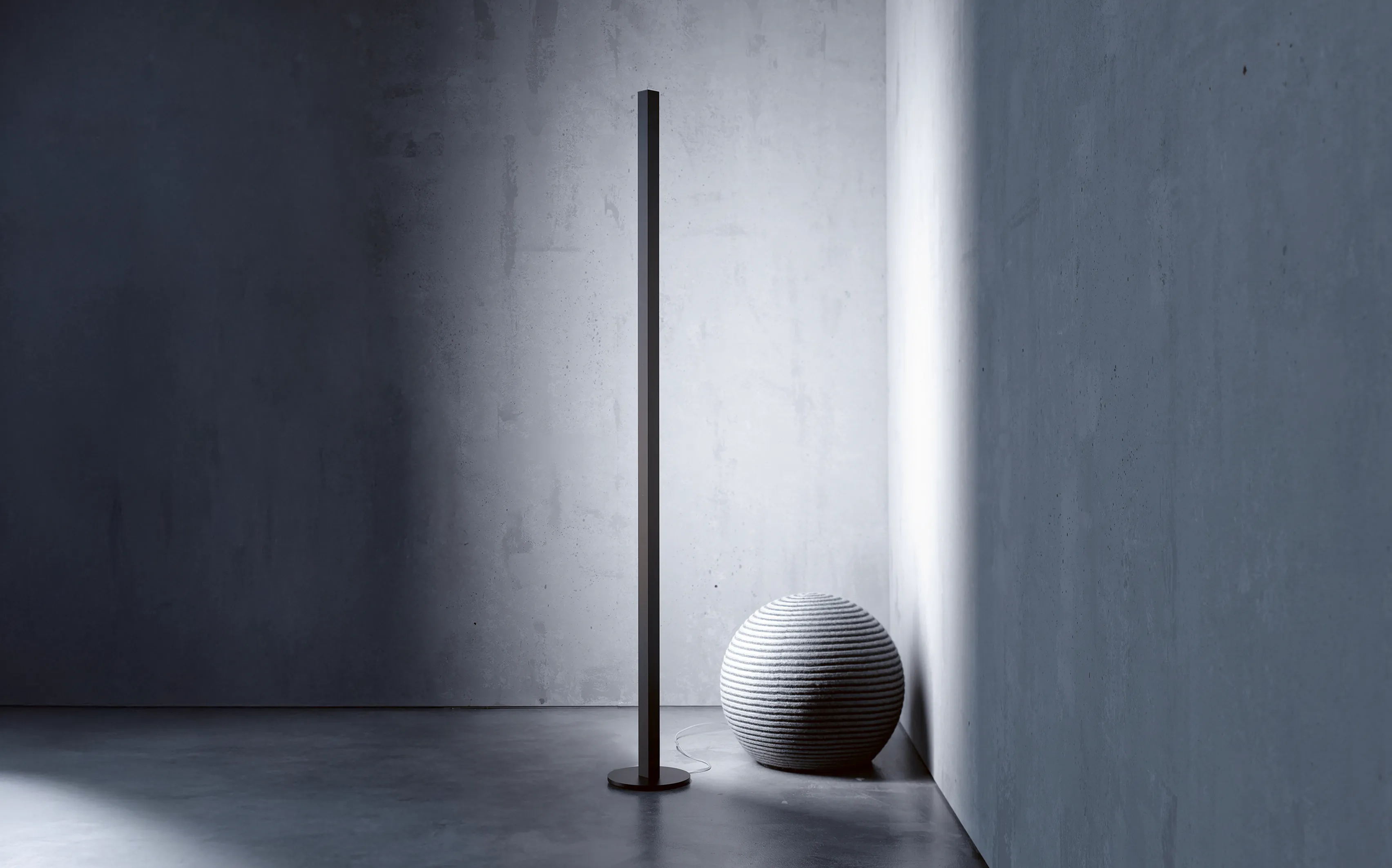 Minimalistische schwarze Stehleuchte GERA Quadra Floor vor einer grauen Betonwand mit rundem, strukturiertem Sitzhocker.