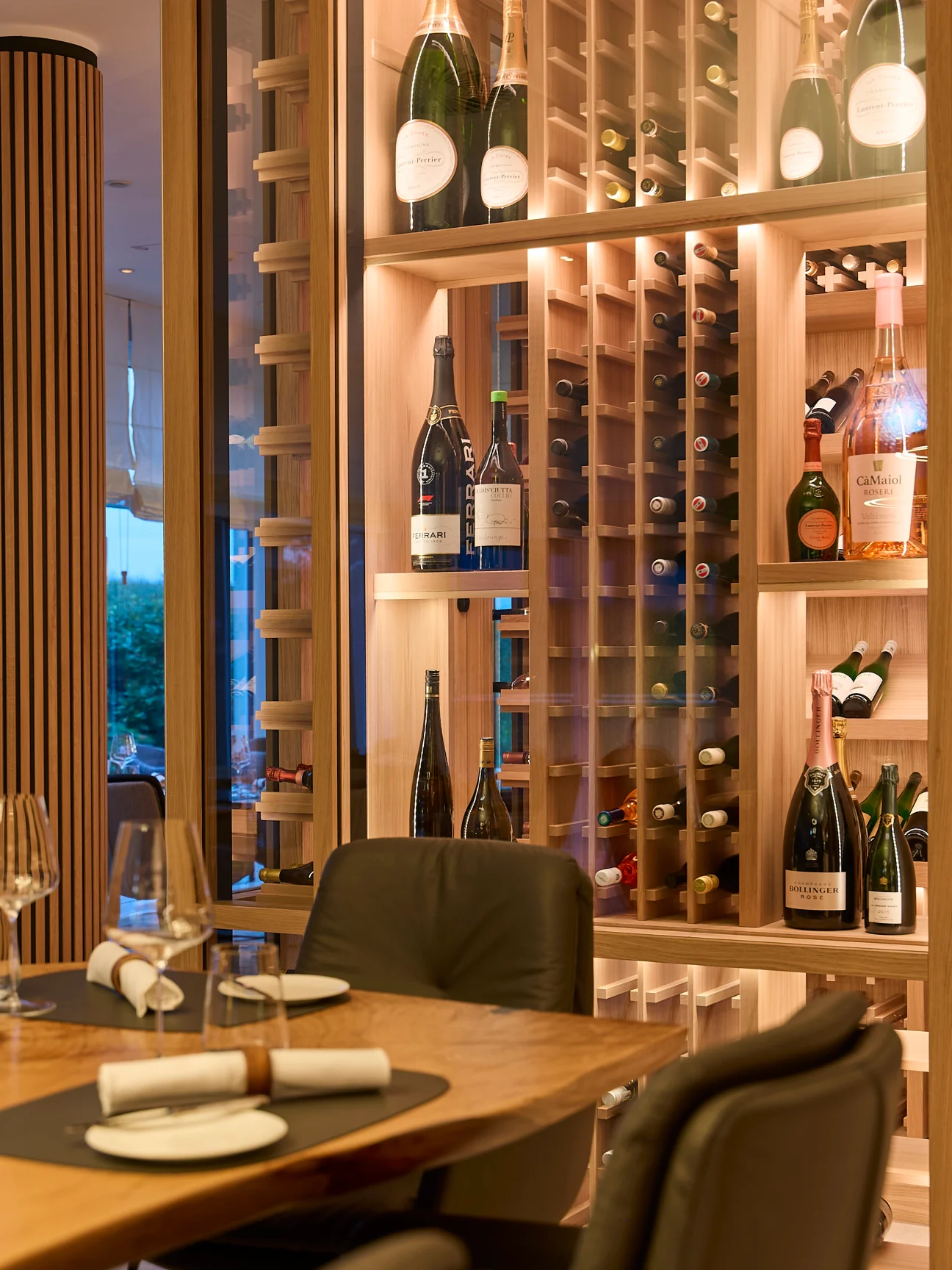 Das Strandhotel Dünenmeer: Ein beleuchteter Weinschrank aus hellem Holz zeigt liegende und stehende Flaschen, darunter Marken wie „FERRARI“ und „BOLLINGER“. Im Vordergrund steht ein gedeckter Holztisch mit Gläsern und weißen Servietten vor dunklen Stühlen. Links geben Holzlamellen den Blick auf die Dämmerung frei.