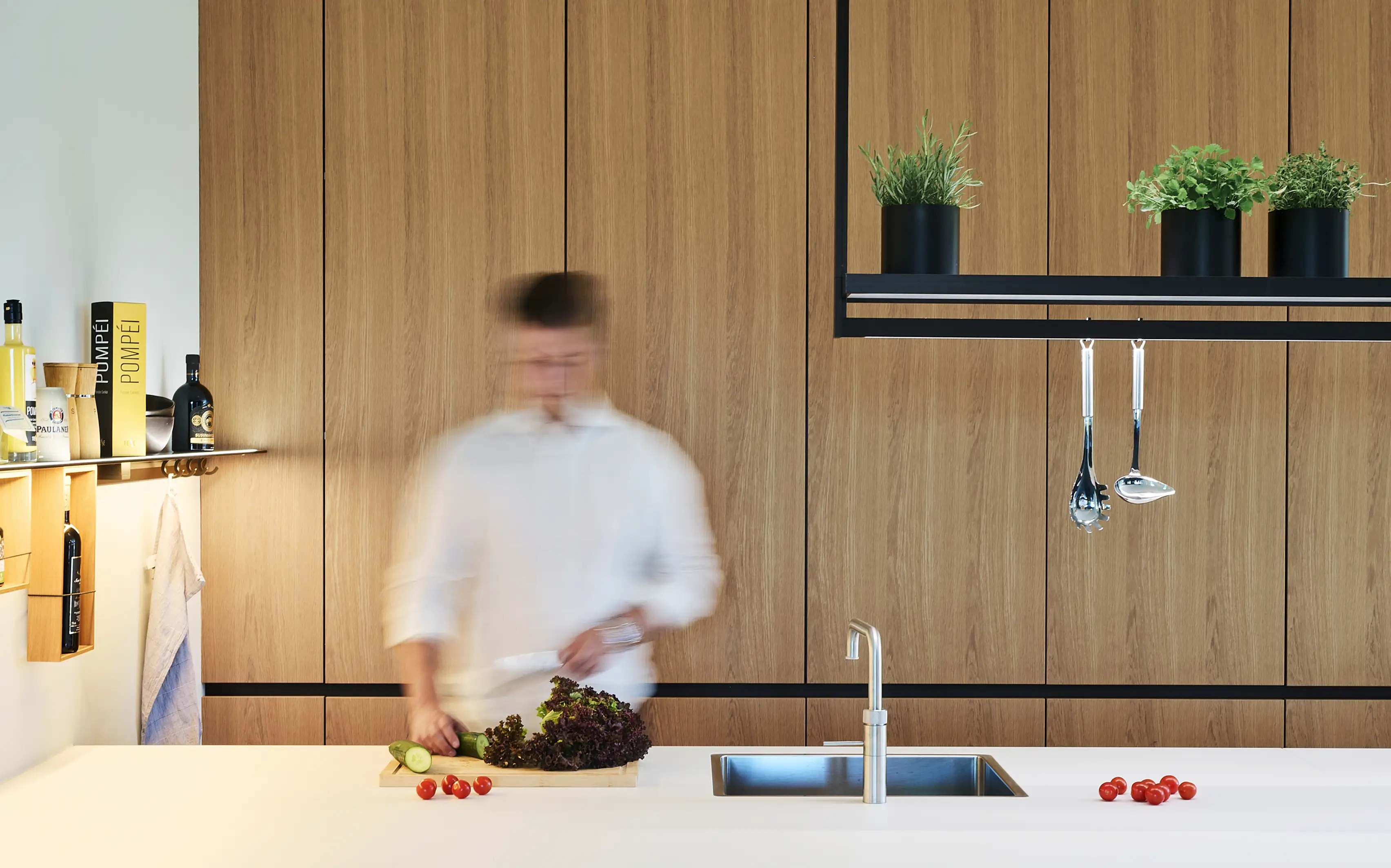 Person bereitet in einer modernen Küche mit Holzschränken Gemüse auf einer weißen Kücheninsel zu. Rechts über ihr ist eine Lichtbrücke GERA Lucina zu sehen.