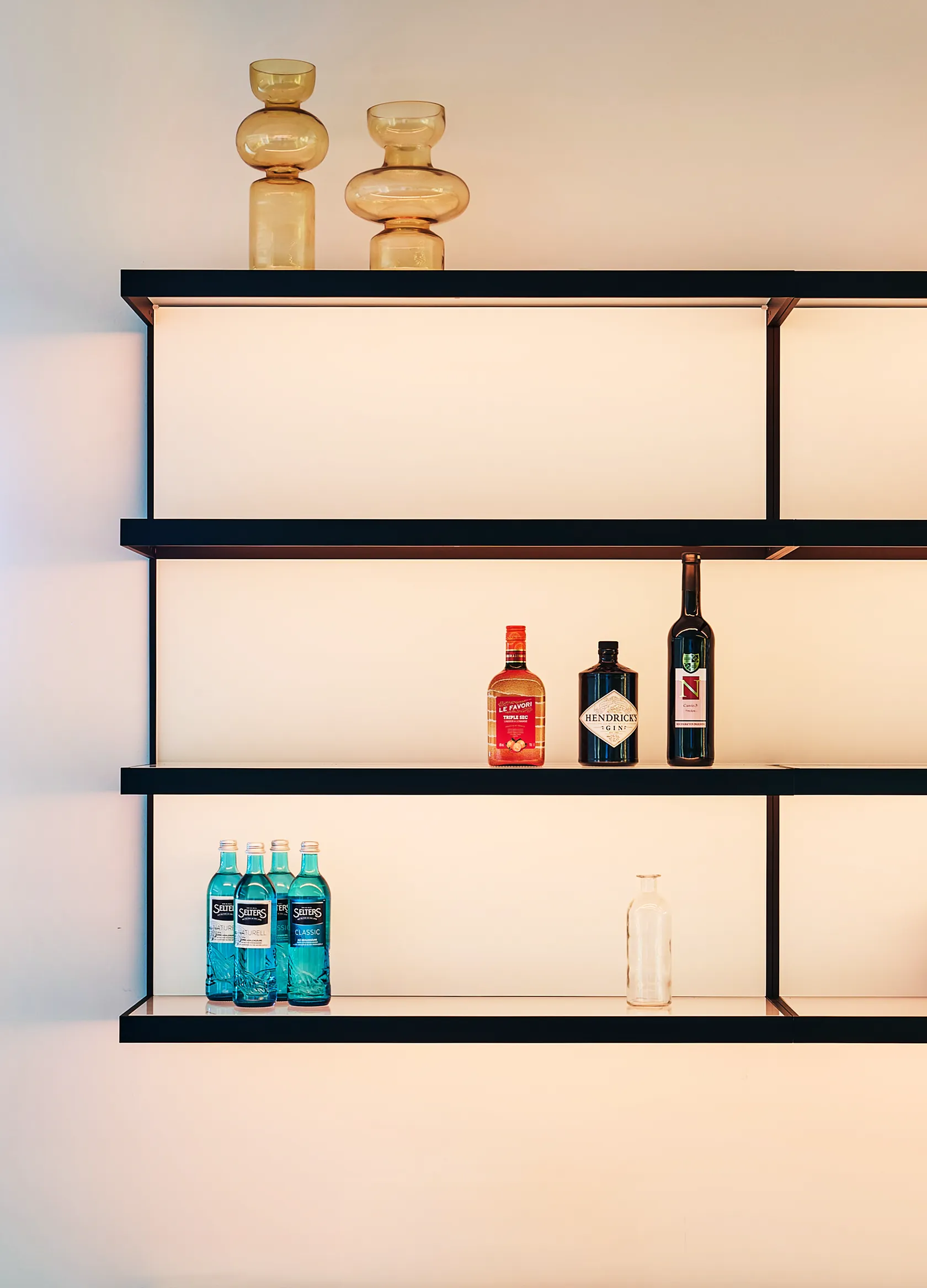 Beleuchtetes GERA Scoop Wandregale mit Glasflaschen, Spirituosen und bernsteinfarbenen Deko-Vasen.