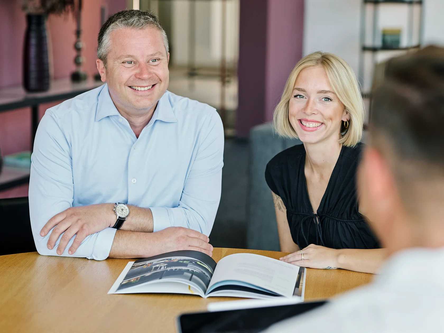 Ein lächelnder Mann im hellblauen Hemd und eine blonde Frau in schwarzer Bluse sitzen an einem runden Holztisch. Sie blicken freundlich zu einer unscharfen Person im Vordergrund. Vor ihnen liegt der aufgeschlagene GERA-Verkaufskatalog.