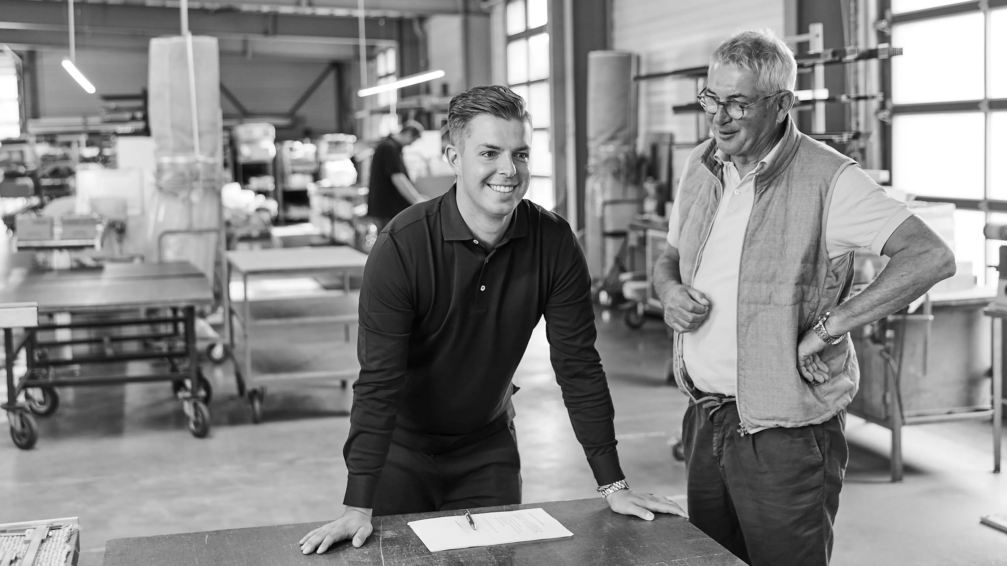 Schwarz-Weiß-Aufnahme in einer Werkhalle von GERA Leuchten: Max Pölitz in dunklem Polohemd stützt sich auf einen Tisch, auf dem ein Dokument mit Stift liegt. Neben ihm steht Geschäftsführer Hening Pölitz, freundlich lächelnd. Der Hintergrund zeigt unscharfe Maschinen und Regale.