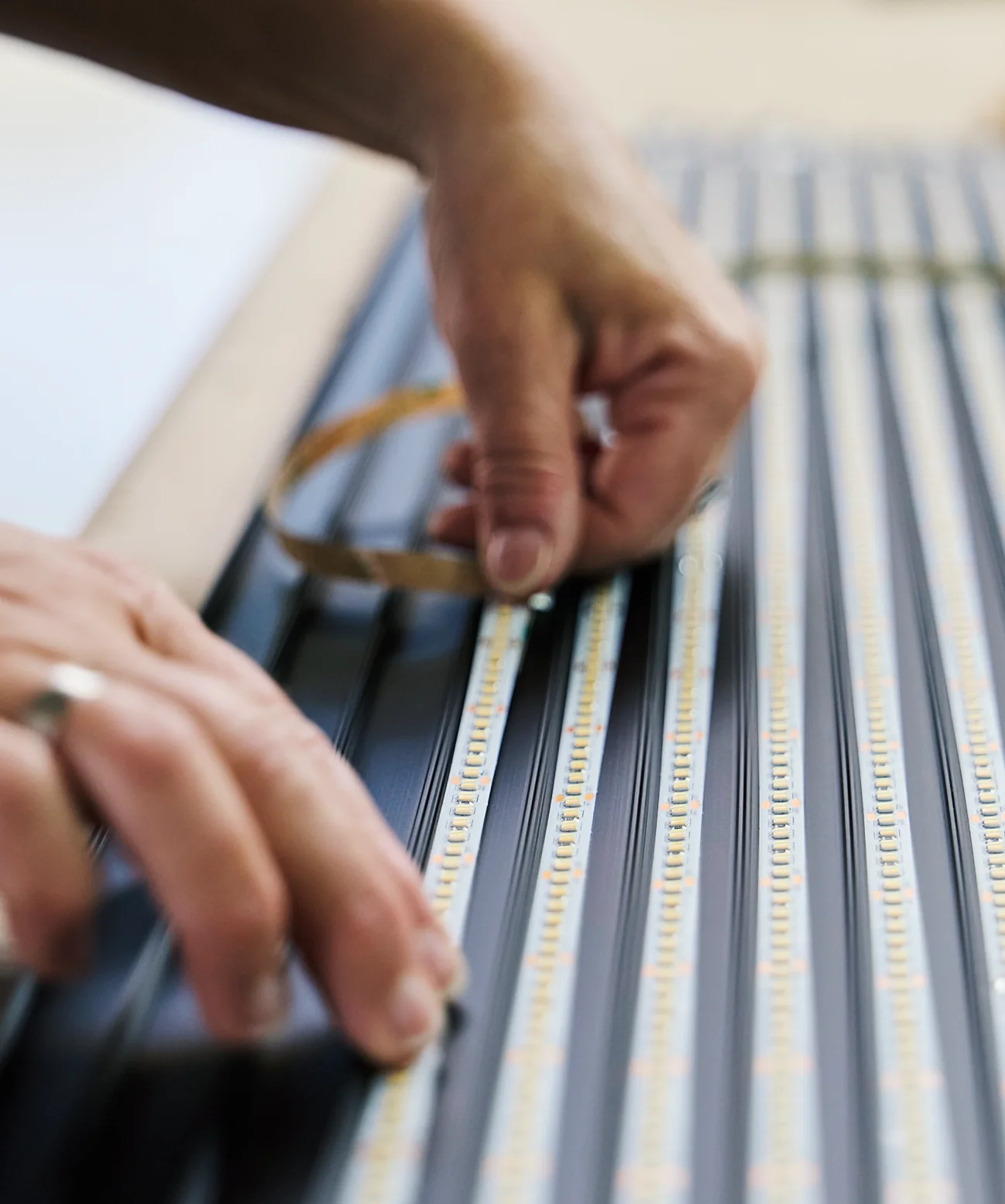 Eine Nahaufnahme zeigt Hände beim präzisen Einsetzen eines LED-Streifens in schwarze Aluminiumprofile. Die rechte Hand zieht das Schutzband ab, während die beringte linke Hand den Streifen fixiert. Mehrere parallele Reihen gelber Leuchtdioden verlaufen diagonal durch das Bild; der Fokus liegt auf der Montage.