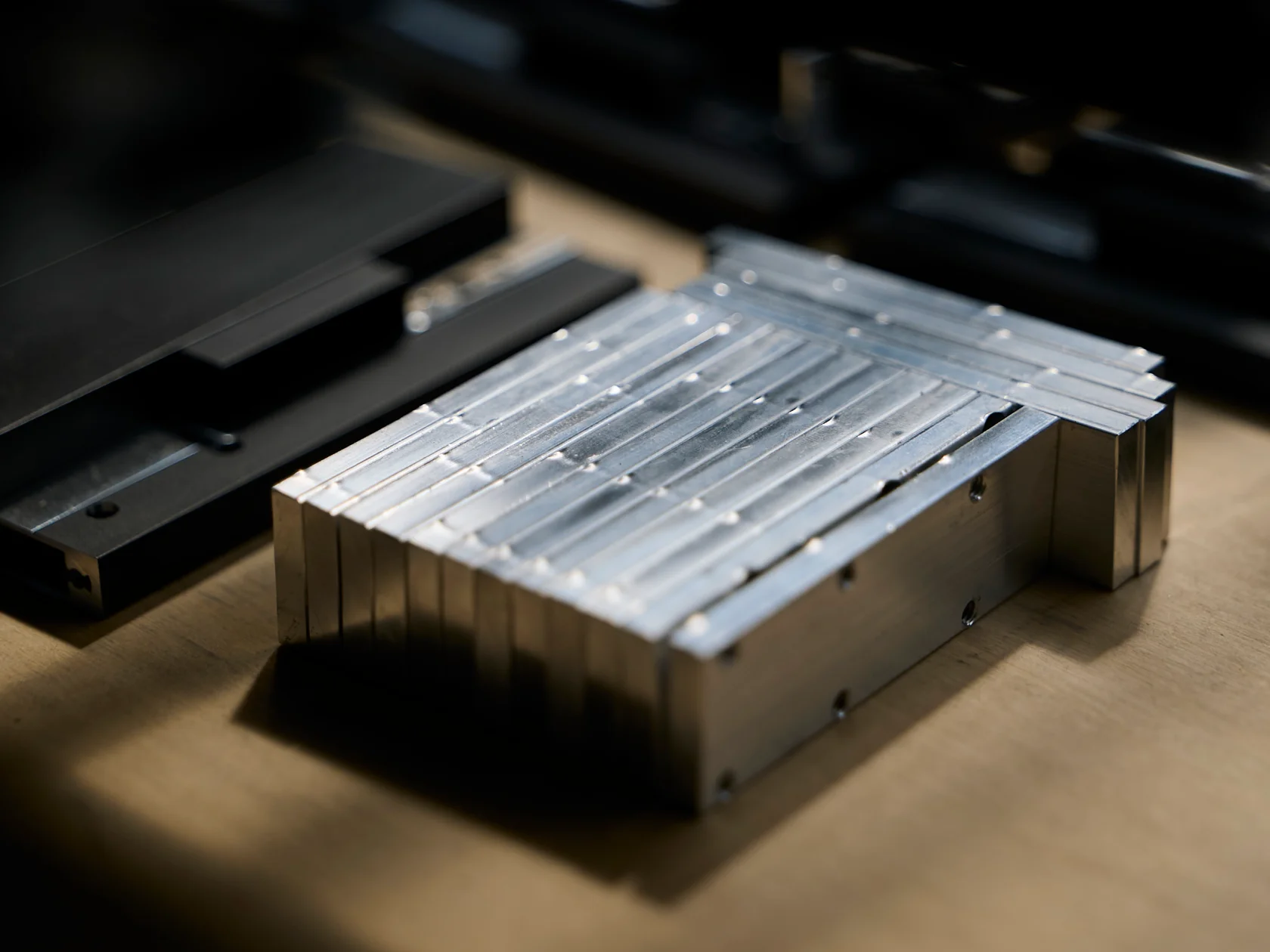 Nahaufnahme eines Stapels gefräster, silberner Metallblöcke auf einer hölzernen Werkbank. Die glänzenden Oberflächen zeigen parallele Rillen, die Seiten Bohrungen. Links liegen unscharfe, schwarze Bauteile.