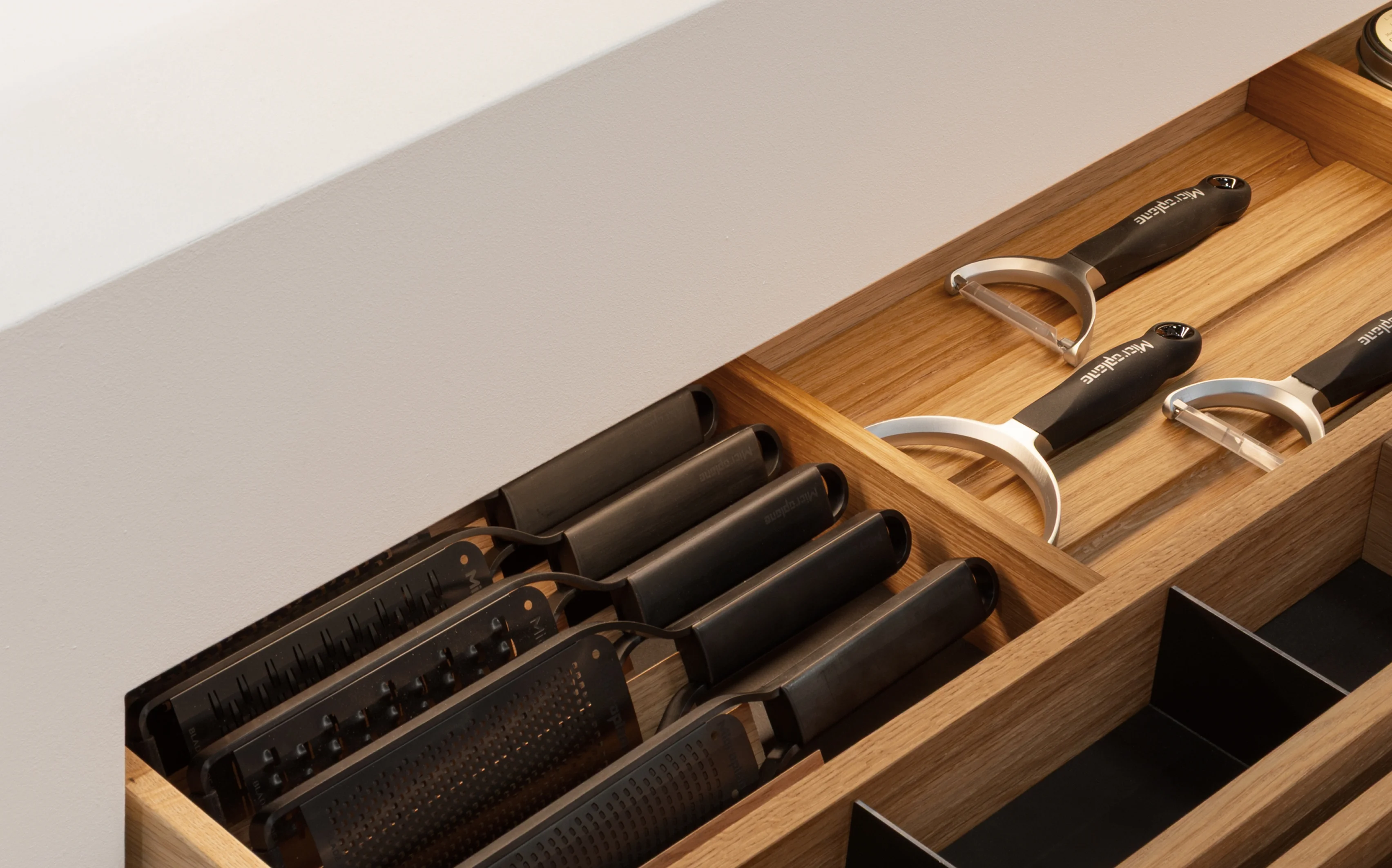 Eine offene hölzerne Küchenschublade mit geordneten Fächern, in denen mehrere Reiben mit schwarzem Griff, Schäler und Küchenutensilien aus Metall aufbewahrt werden. Von oben sanft beleuchtet von der GERA Auszugbeleuchtung.