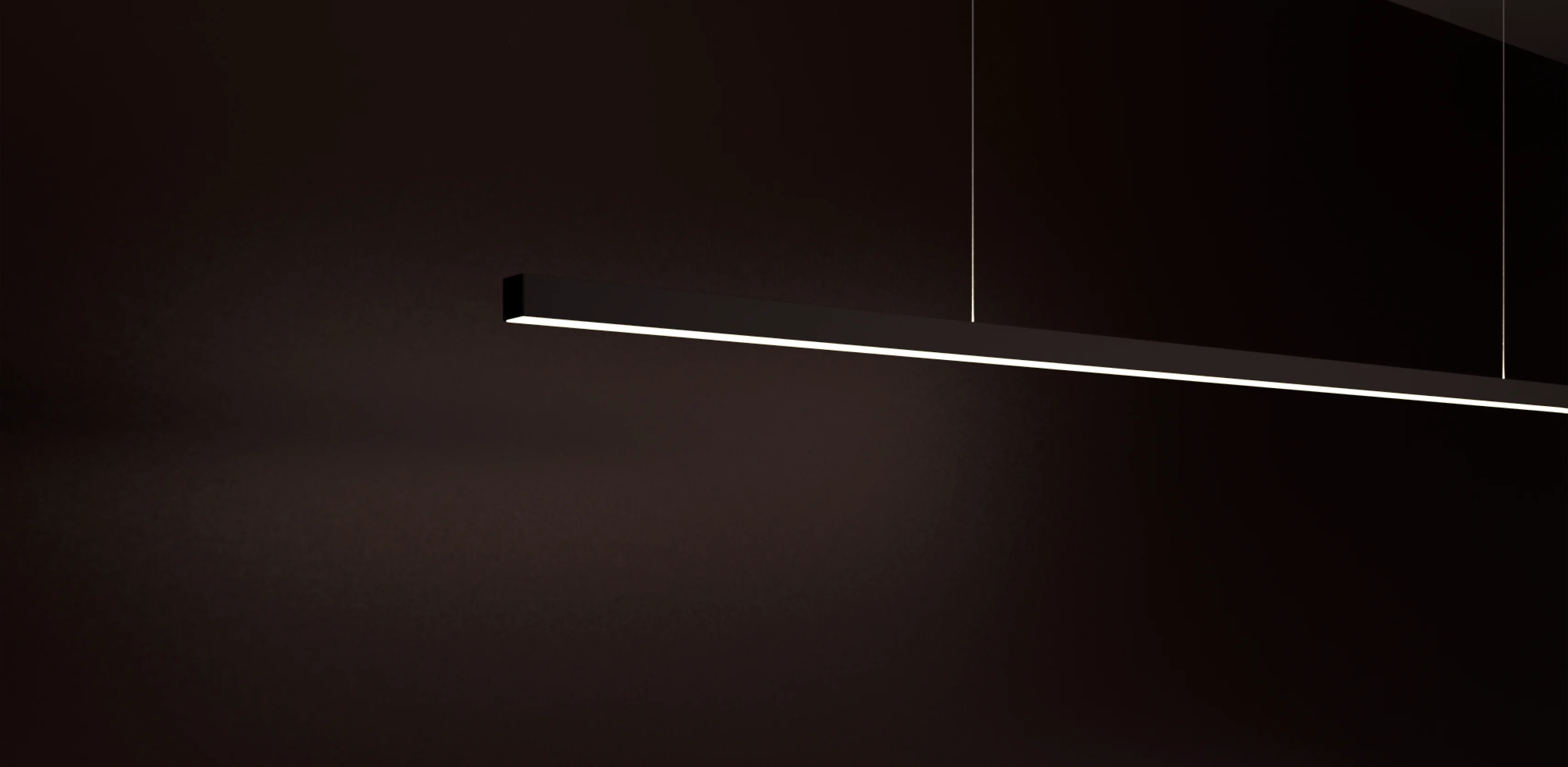 Vor einem tief dunkelbraunen Hintergrund hängt eine minimalistische, lineare Pendelleuchte, GERA Quadra Swing. Das lange, schwarze, quadratische Aluminiumprofil schwebt schräg im Raum, gehalten von filigranen stromführenden Drähten. Das integrierte weiße LED-Lichtband an der Unterseite strahlt hell und bildet einen starken Kontrast zur düsteren Umgebung.
