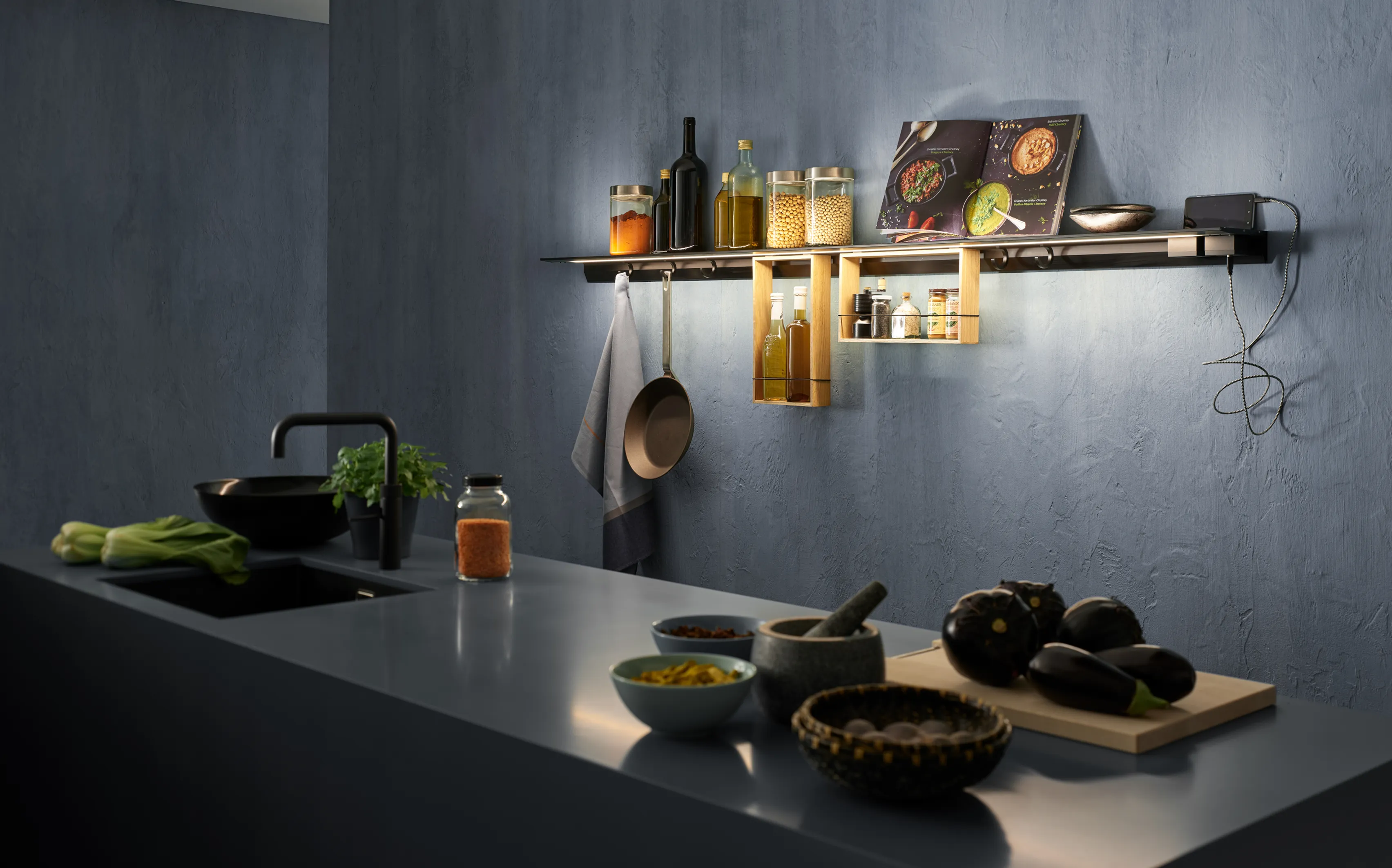 Moderne Küchenzeile mit beleuchtetem Lichtbord GERA Feature, Kochutensilien und Zutaten auf einer dunklen Arbeitsplatte.