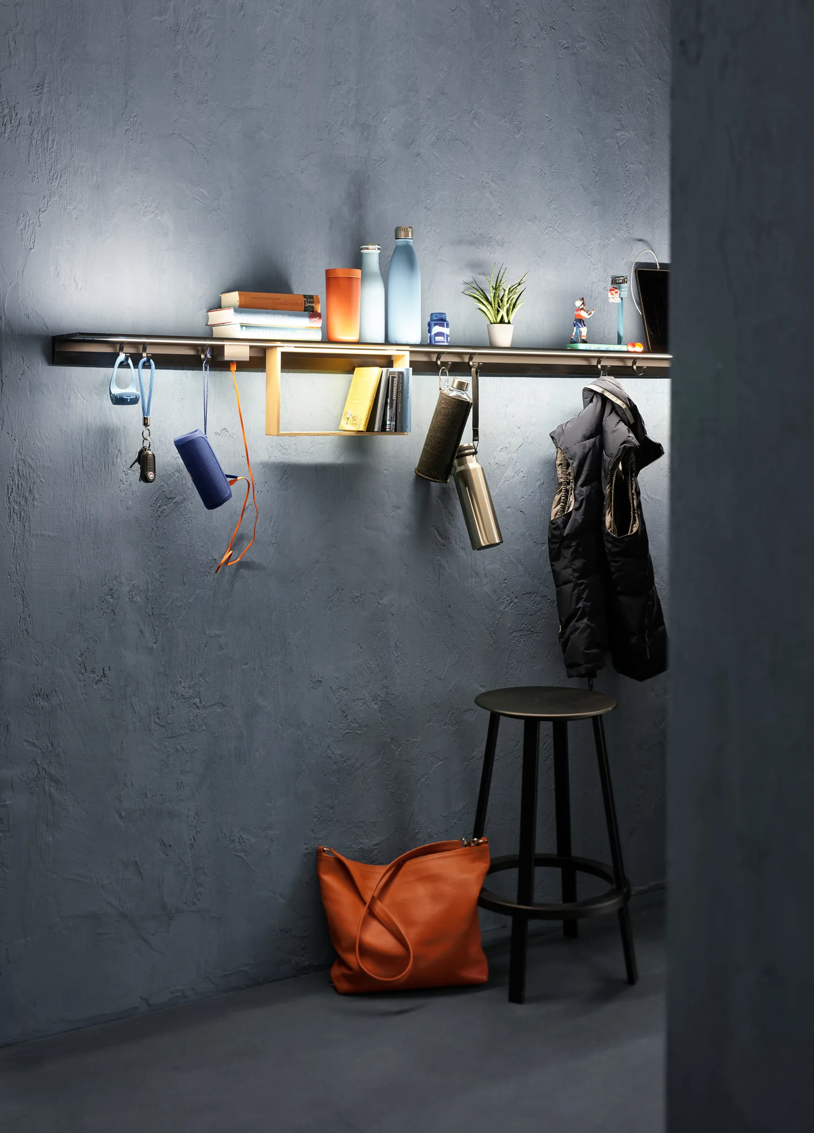 Beleuchtetes Lichtbord GERA Feature mit Haken, daran hängenden Accessoires, Büchern, Flaschen und einer Weste, ergänzt durch einen Hocker und eine Tasche.