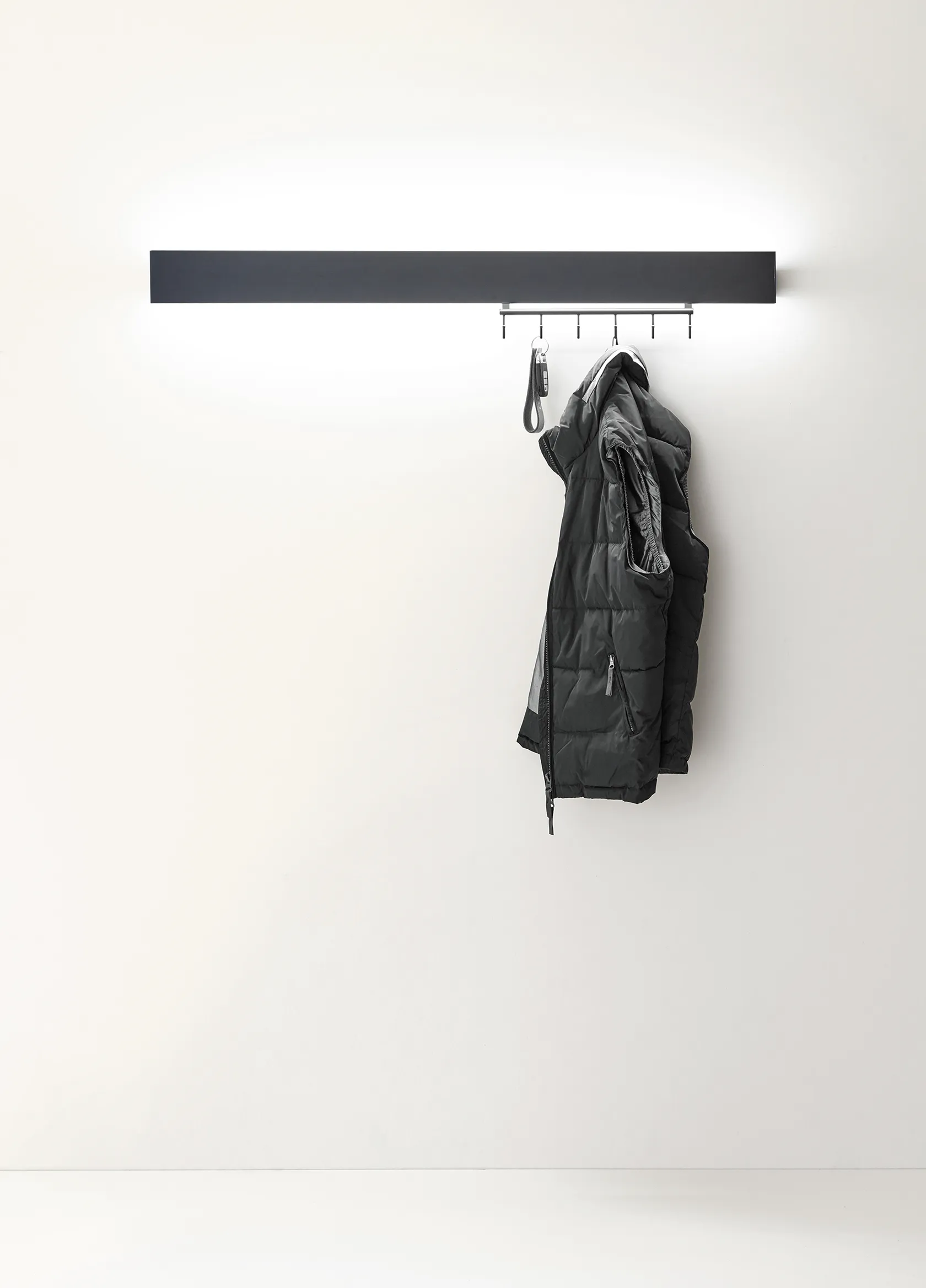 Beleuchtete schwarze Garderobenleuchte GERA Hook mit einer hängenden Steppweste vor weißer Wand.