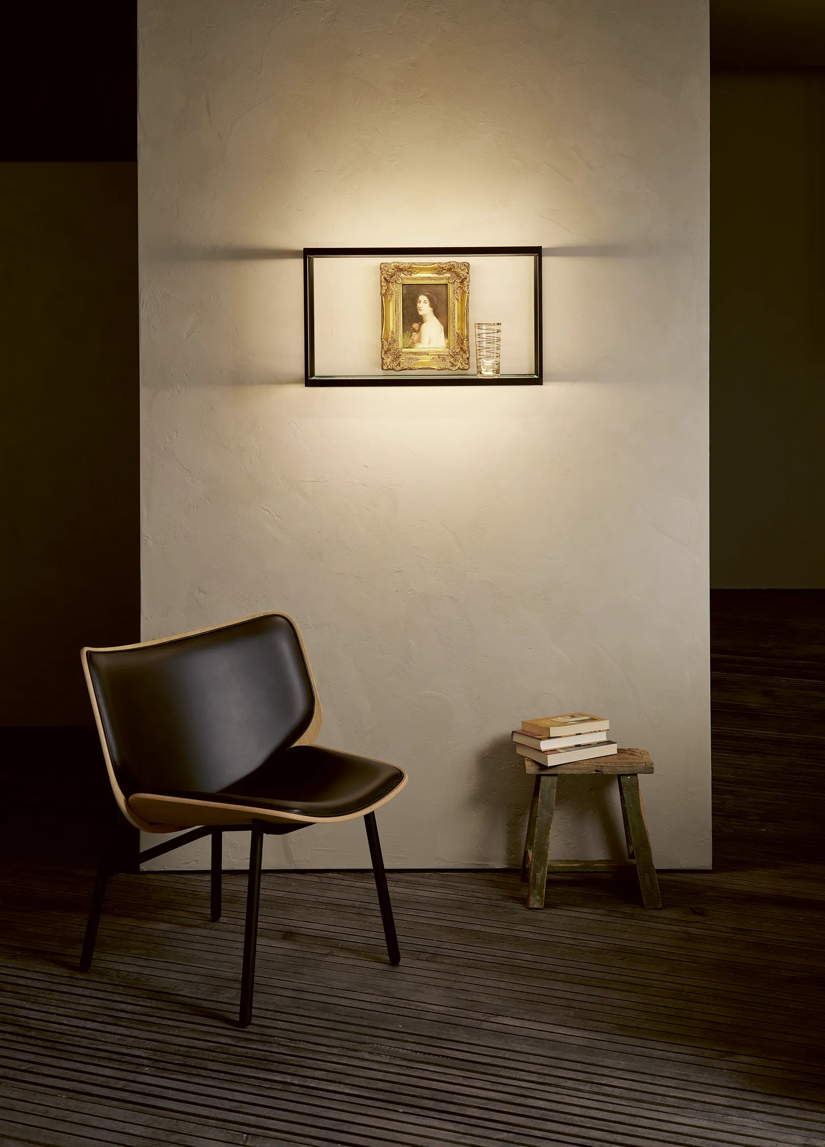 Ein moderner schwarzer Stuhl steht vor einer hellen, strukturierten Wand. Darüber ruhen ein gerahmtes Kunstwerk und ein Glas in einem sanft beleuchteten GERA Cube. Neben dem Stuhl steht ein kleiner Holzhocker mit einem Stapel Bücher.