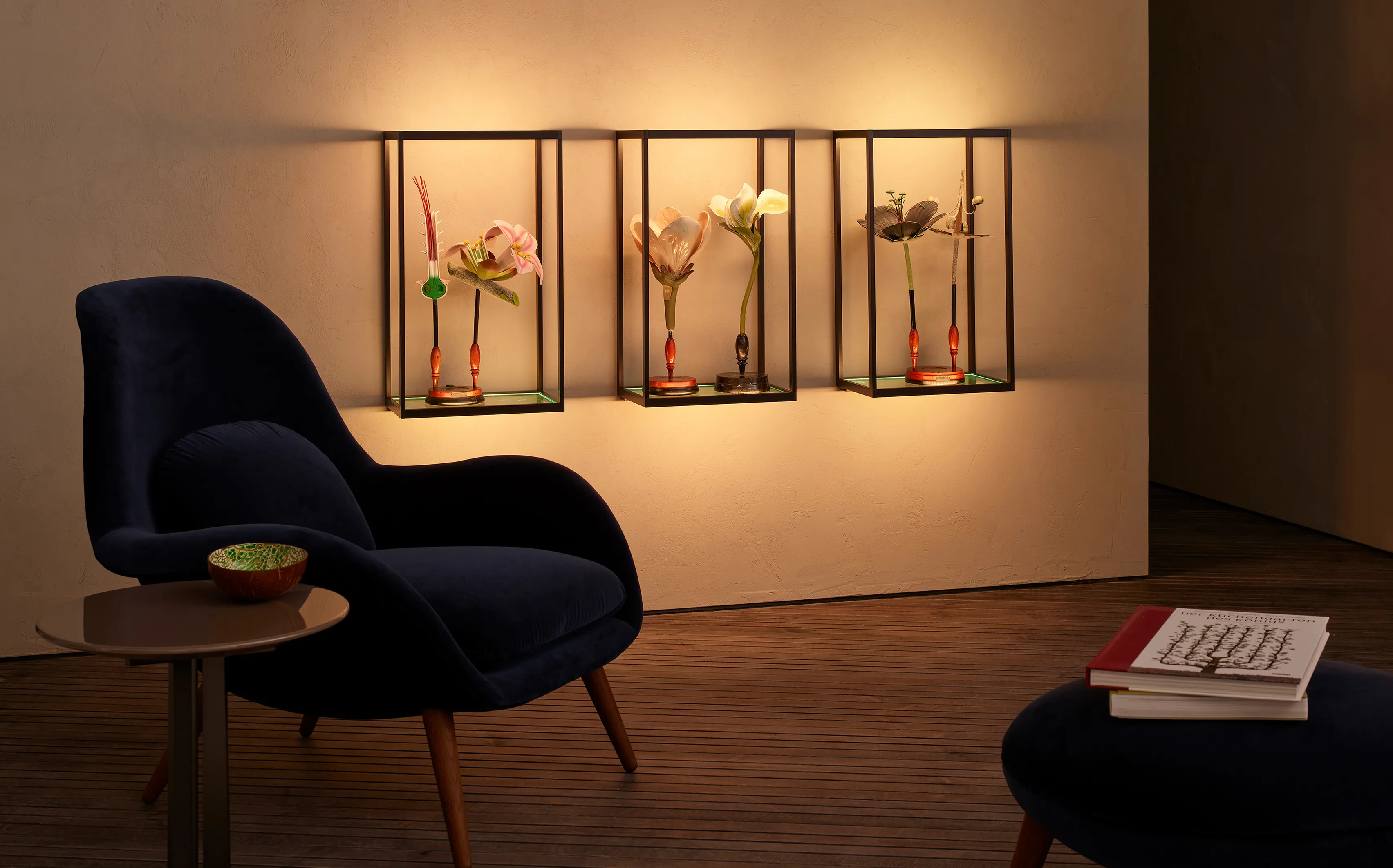 Ein gemütlicher Raum mit einem dunkelblauen Sessel, einem kleinen runden Tisch und einem Ottomanen. Drei an der Wand befestigte, warm beleuchtete GERA Cubes rahmen exotische Blumen-Skulpturen gekonnt ein.