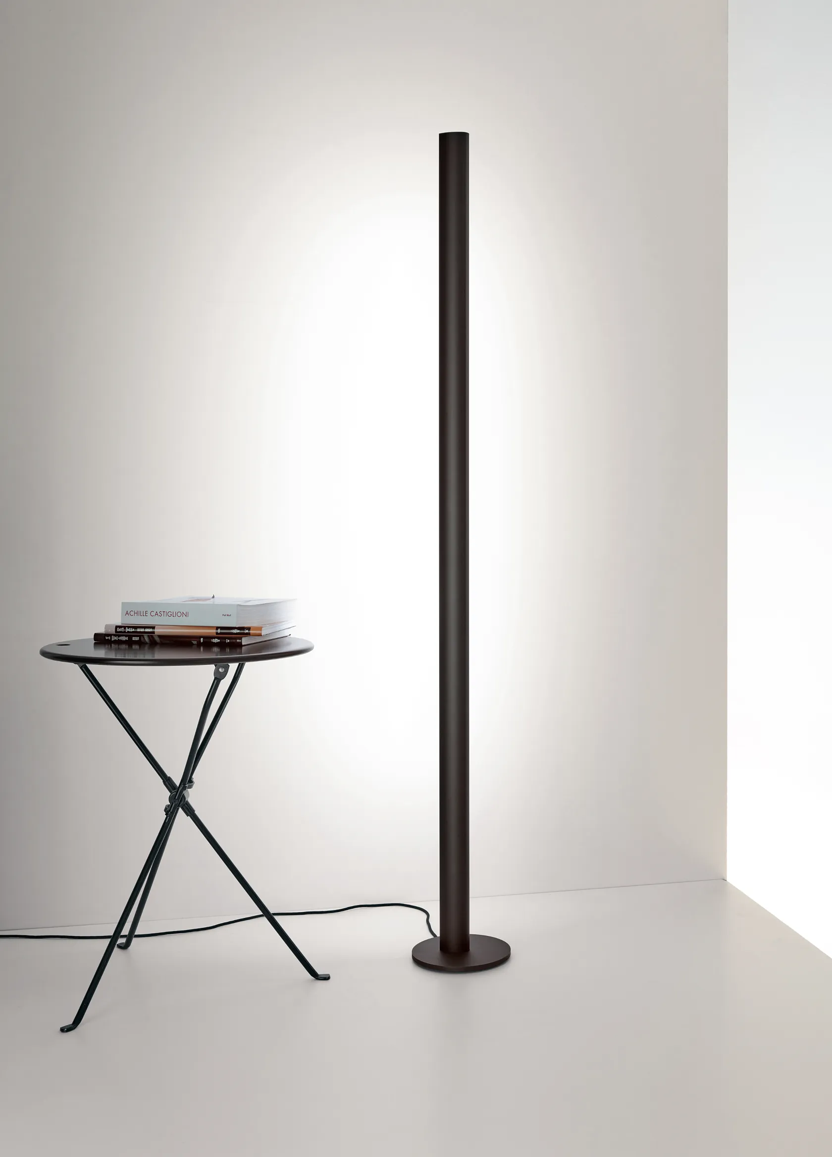 Die schlanke Stehleuchte GERA Avion wirft sanftes Licht auf eine weiße Wand neben einem kleinen runden Tisch mit gestapelten Büchern in einem minimalistischen Raum.