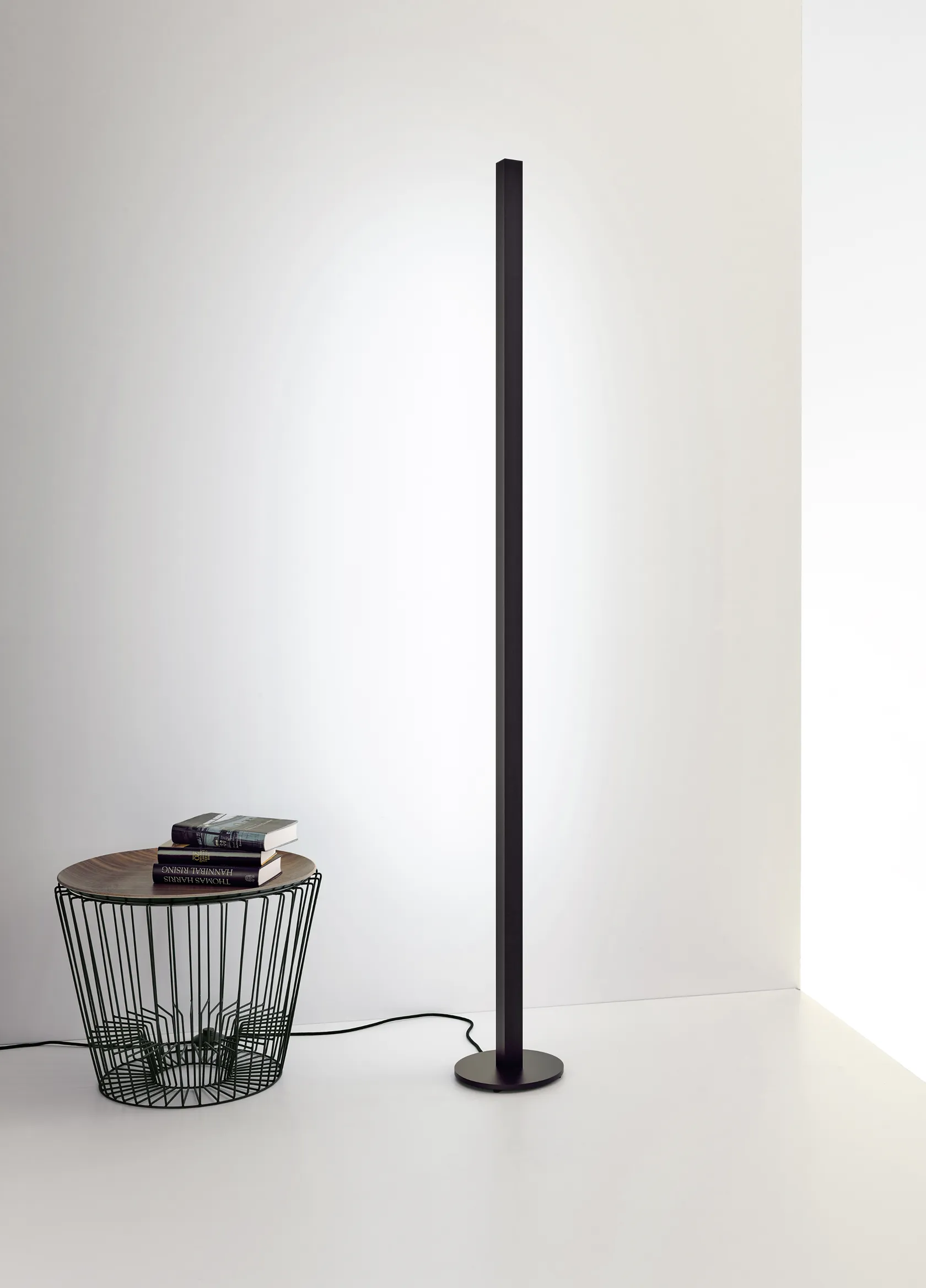 Minimalistische schwarze Stehleuchte GERA Quadra Floor neben einem Beistelltisch aus Draht mit Büchern.