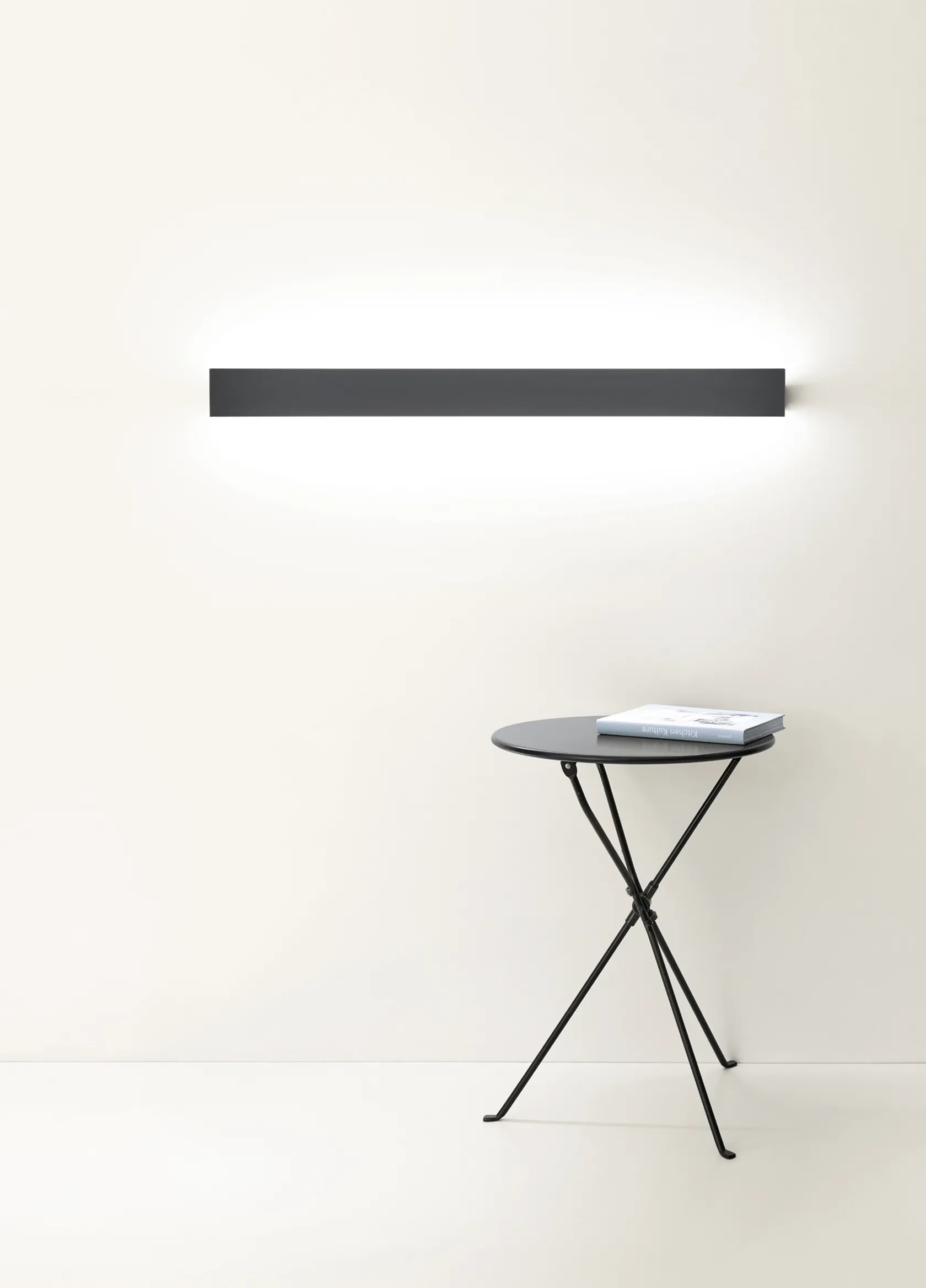 Schwarze Wandleuchte GERA Slant über einem kleinen runden Beistelltisch mit Buch vor weißer Wand.