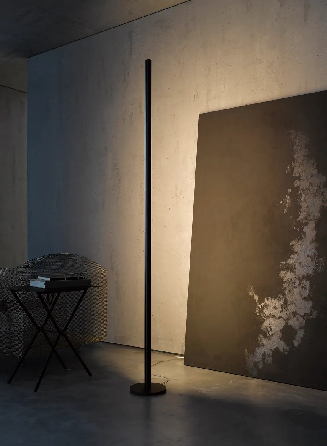 Ein moderner Betonraum mit einer hohen schwarzen Stehleuchte GERA Avion, die ein großes abstraktes Bild beleuchtet. Links steht ein aus Metall geflochtener Sessel neben einem kleinen Tisch mit Büchern.
