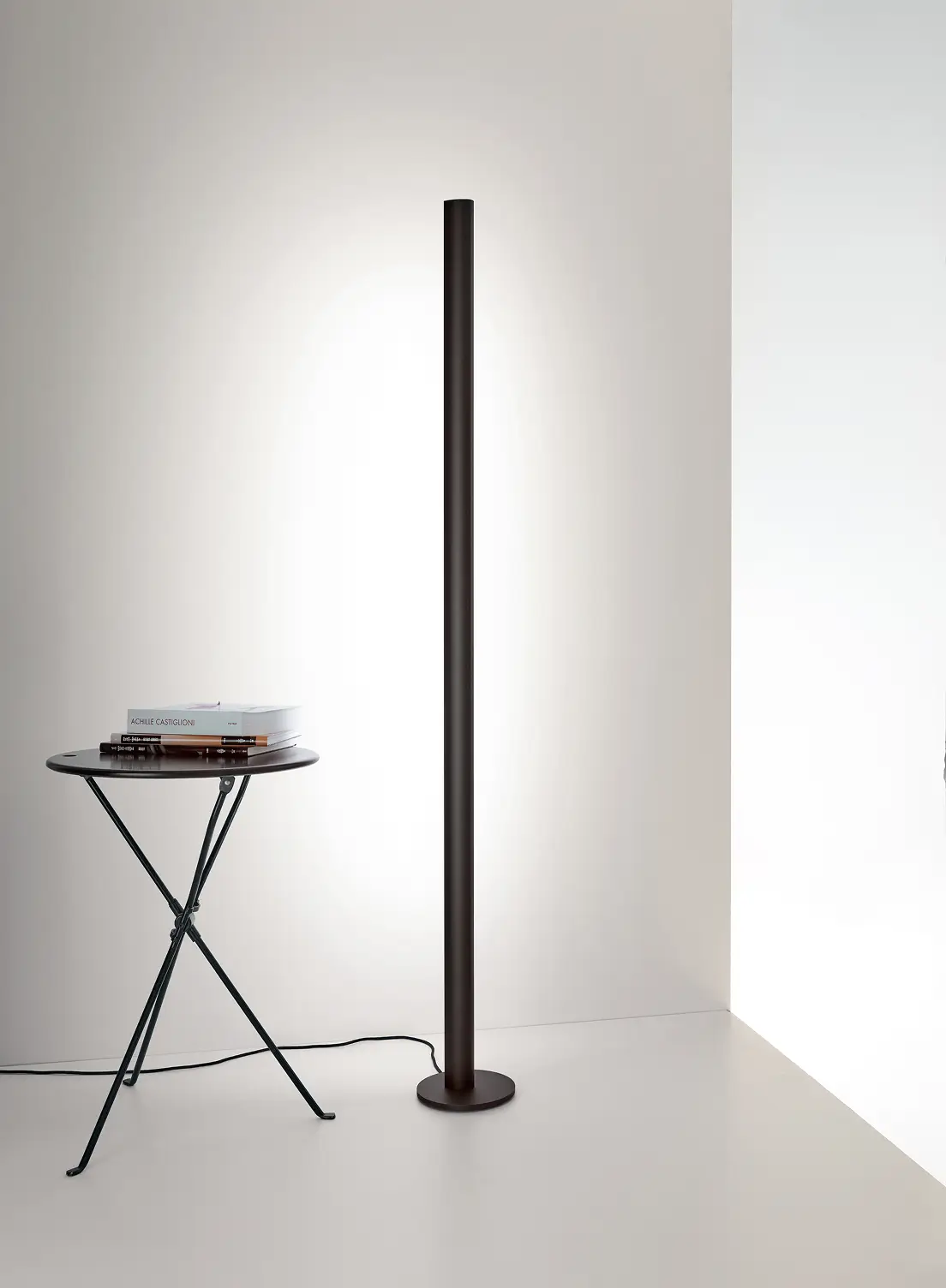 Schlanke Stehleuchte GERA Avion neben einem kleinen Beistelltisch mit Büchern vor einer hellen Wand.