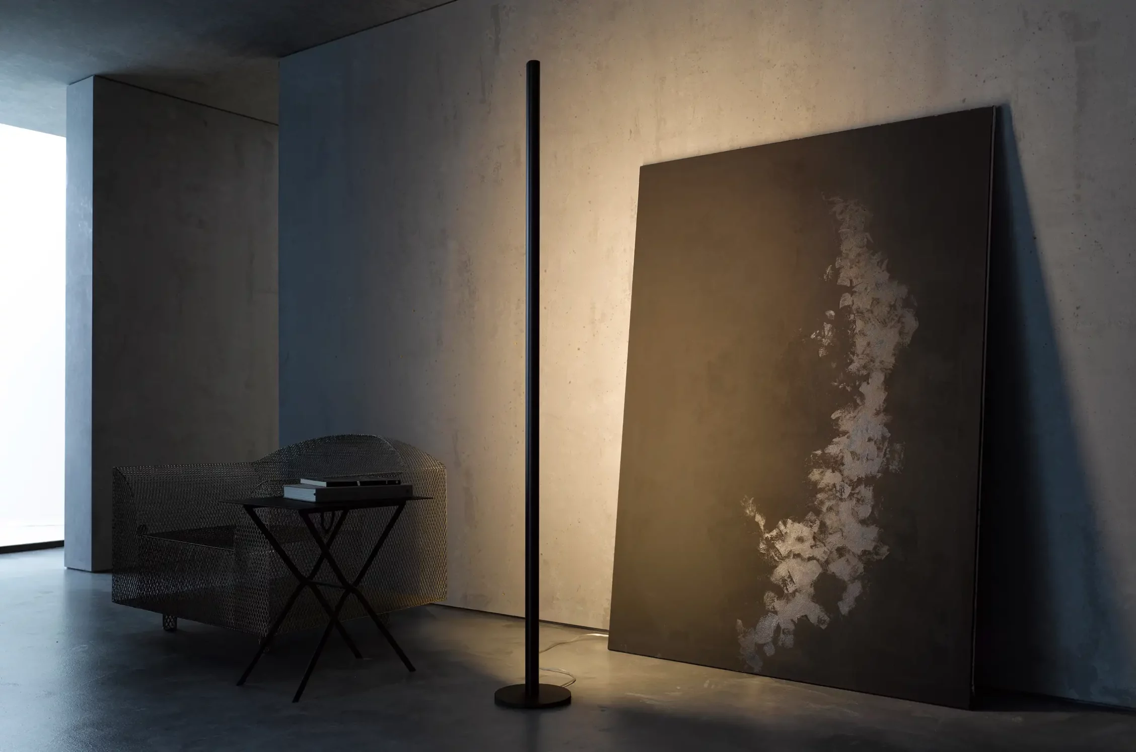 Ein moderner Betonraum mit einer hohen schwarzen Stehleuchte GERA Avion, die ein großes abstraktes Bild beleuchtet. Links steht ein aus Metall geflochtener Sessel neben einem kleinen Tisch mit Büchern.