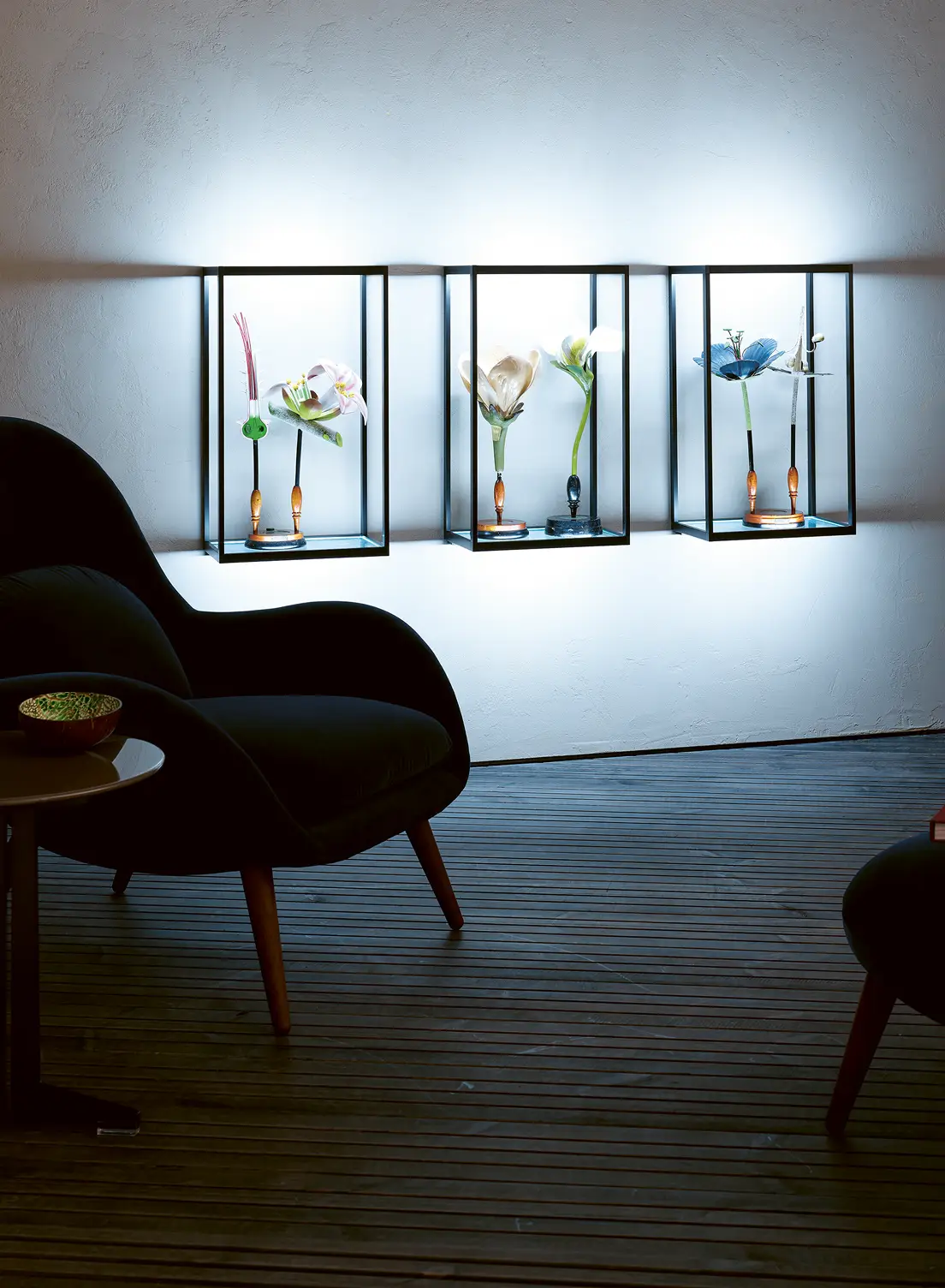 Drei beleuchtete GERA Cube Wandregale mit floralen Objekten über einem schwarzen Sessel in einem modernen Raum.