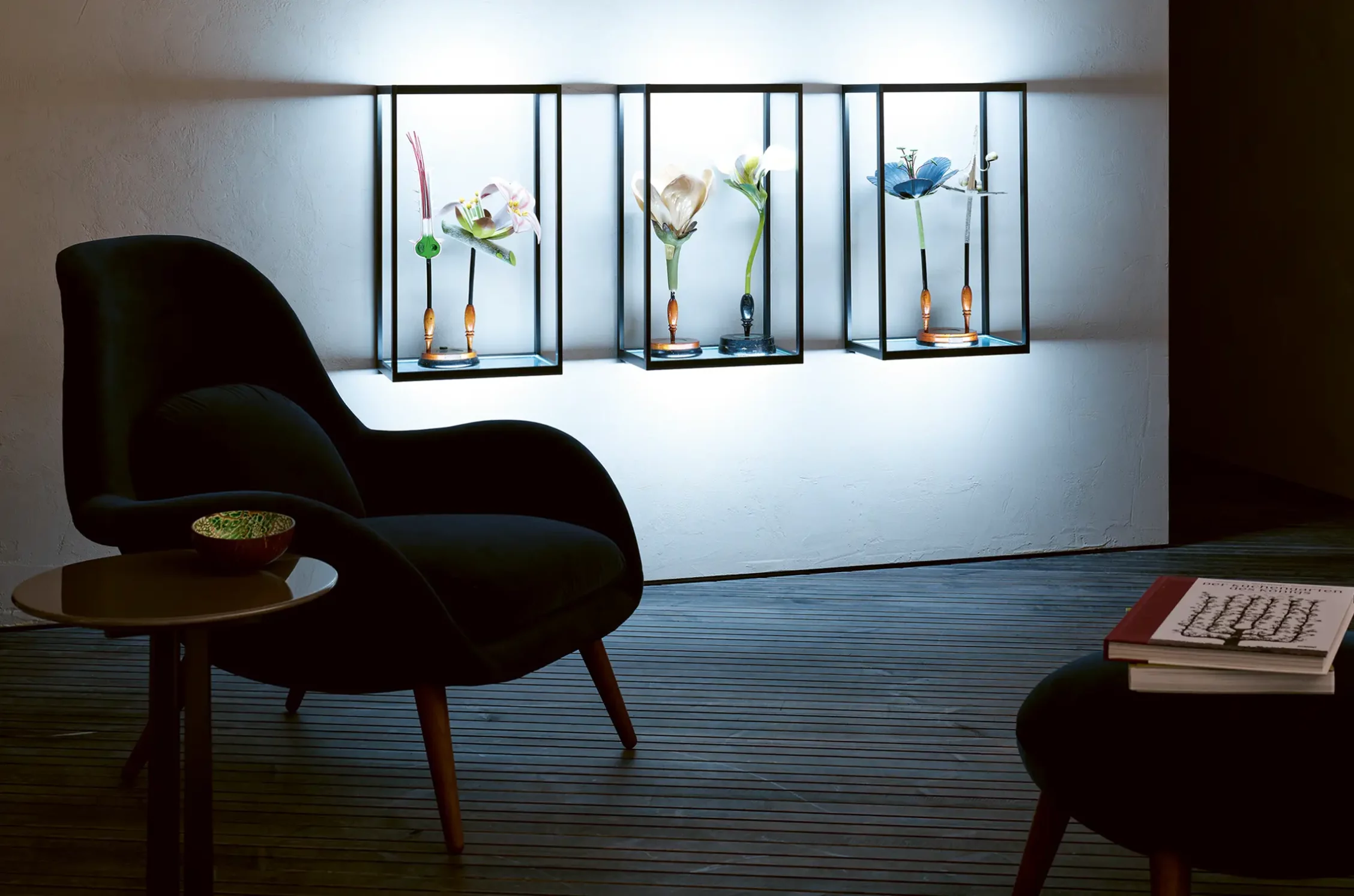 Drei beleuchtete GERA Cube Wandregale mit floralen Objekten über einem schwarzen Sessel in einem modernen Raum.
