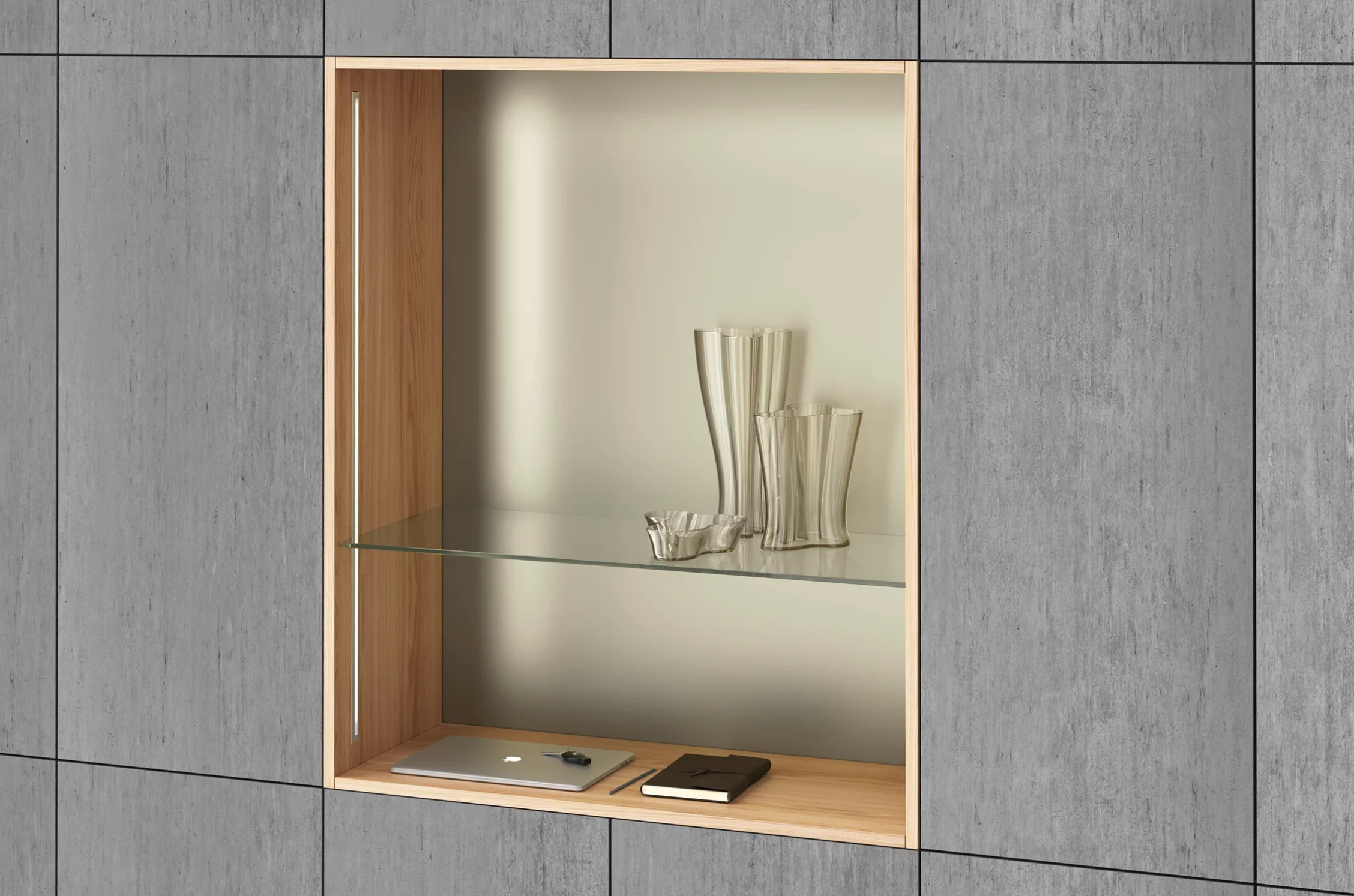 Ein modernes Einbauregal umgeben von grauen Wandpanelen. Darin der drei dekorative Glasvasen, ein zugeklappter Laptop und ein Notizbuch im Ledereinband. Beleuchtet wird das Regal durch vertikal eingesetzte GERA Einbauleuchten indirekt.