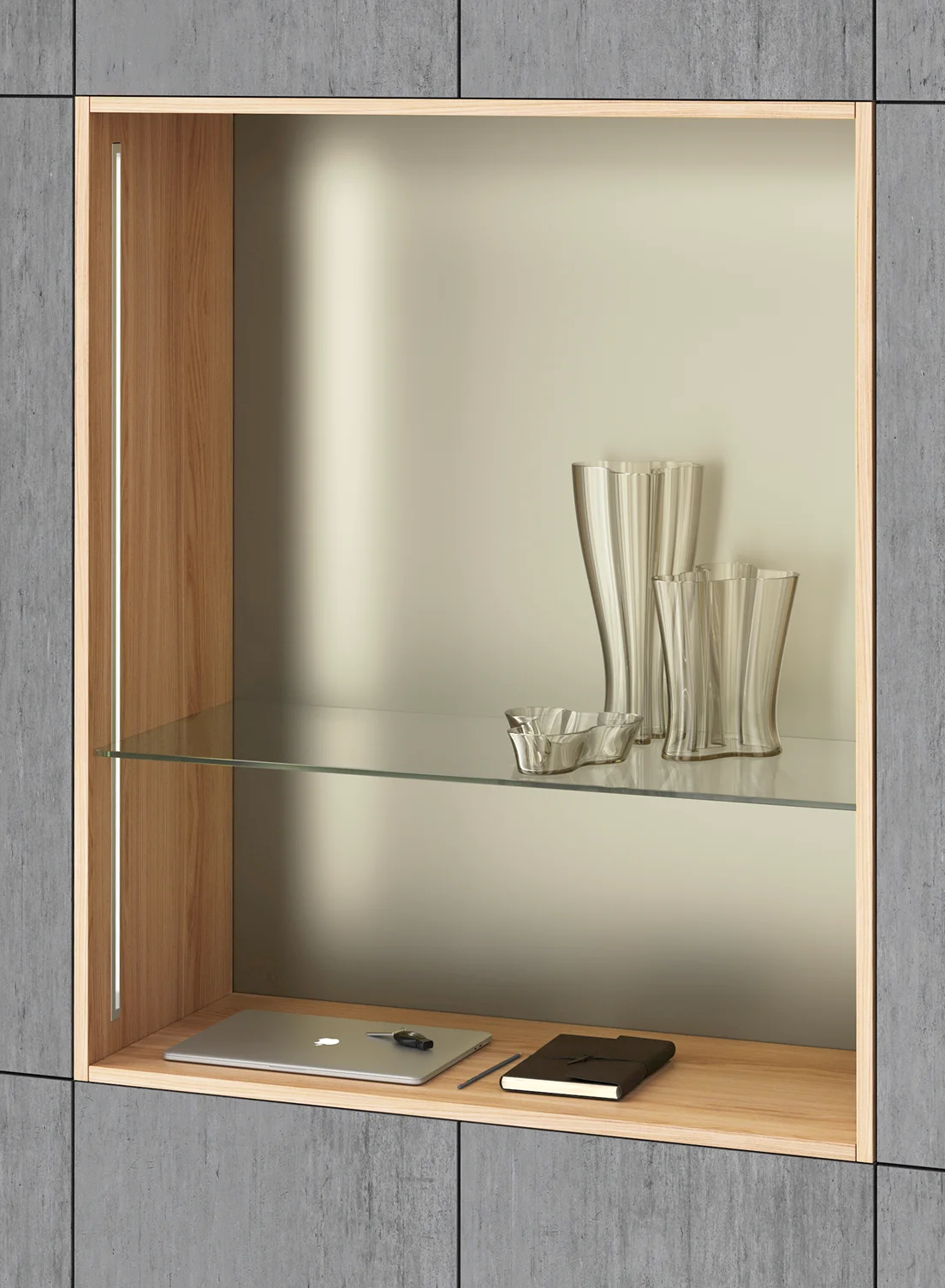 Ein modernes Einbauregal umgeben von grauen Wandpanelen. Darin der drei dekorative Glasvasen, ein zugeklappter Laptop und ein Notizbuch im Ledereinband. Beleuchtet wird das Regal durch vertikal eingesetzte GERA Einbauleuchten indirekt.