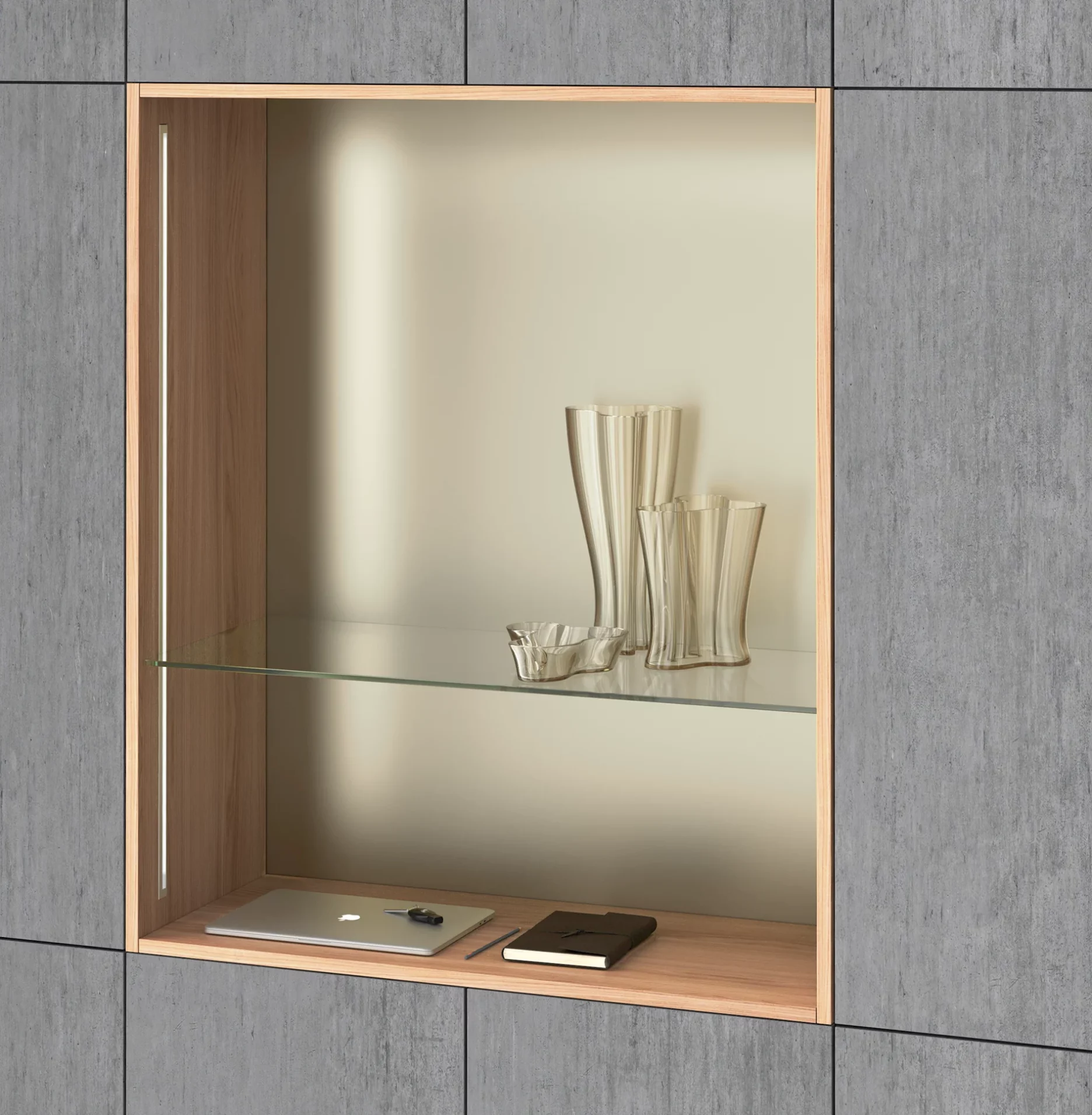 Ein modernes Einbauregal umgeben von grauen Wandpanelen. Darin der drei dekorative Glasvasen, ein zugeklappter Laptop und ein Notizbuch im Ledereinband. Beleuchtet wird das Regal durch vertikal eingesetzte GERA Einbauleuchten indirekt.