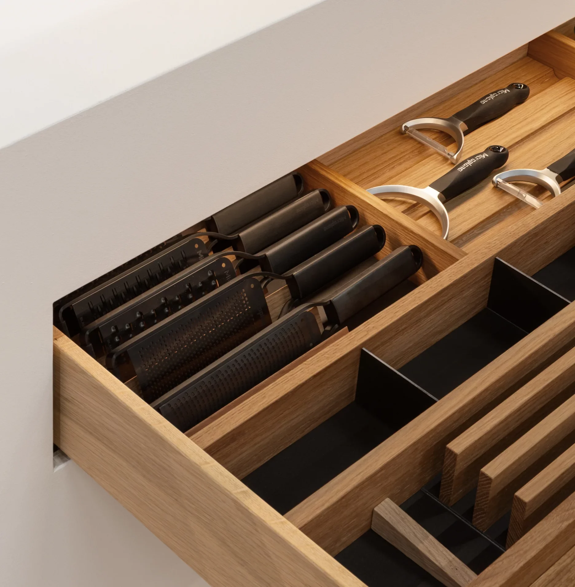 Eine offene hölzerne Küchenschublade mit geordneten Fächern, in denen mehrere Reiben mit schwarzem Griff, Schäler und Küchenutensilien aus Metall aufbewahrt werden. Von oben sanft beleuchtet durch die GERA Auszugbeleuchtung.