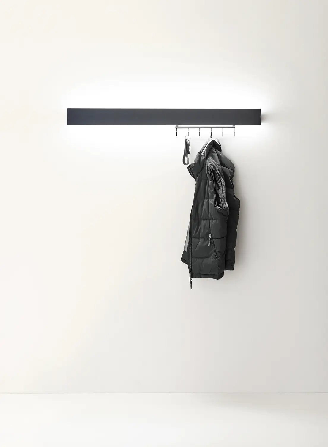 Beleuchtete schwarze Garderobenleuchte GERA Hook mit einer hängenden Steppweste vor weißer Wand.