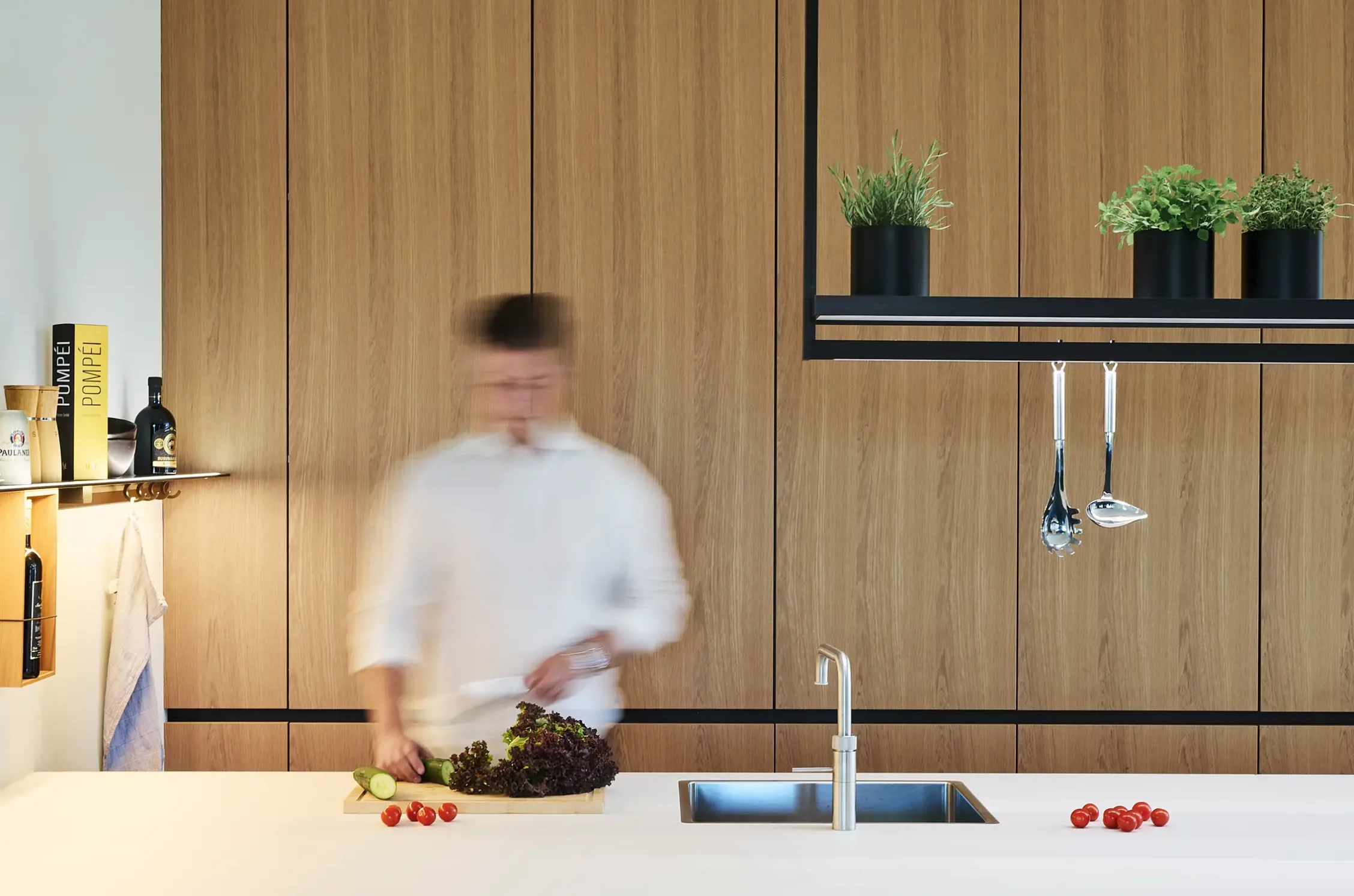 Person bereitet in einer modernen Küche mit Holzschränken Gemüse auf einer weißen Kücheninsel zu. Rechts über ihr ist eine Lichtbrücke GERA Lucina zu sehen.
