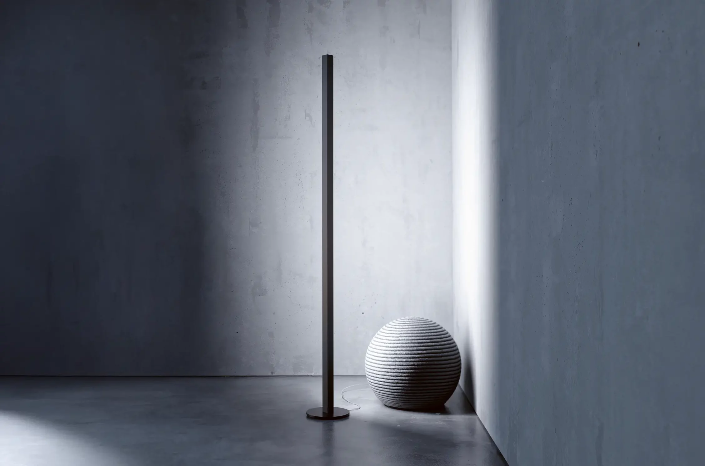 Schlanke schwarze Stehleuchte GERA Quadra Floor neben einem strukturierten, kugelförmigen Sitzmöbel in einer minimalistischen Raumecke.