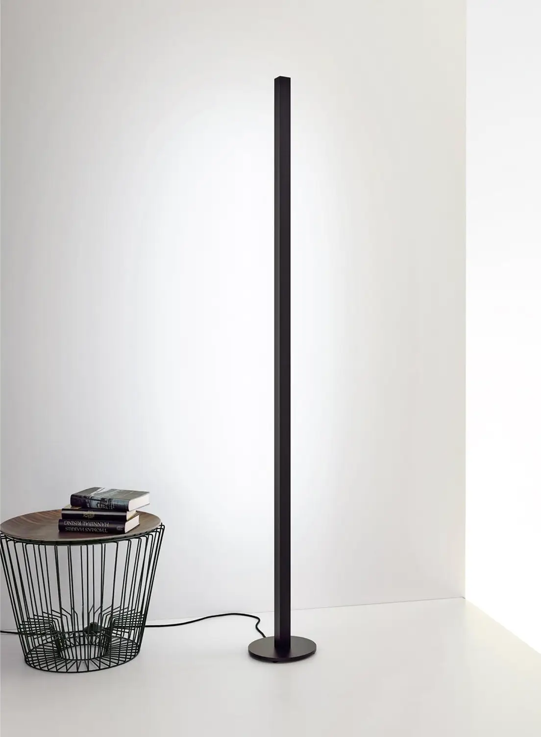 Schlanke schwarze Stehleuchte GERA Quadra Floor neben einem kleinen Drahtkorb-Beistelltisch mit Büchern vor einer weißen Wand.
