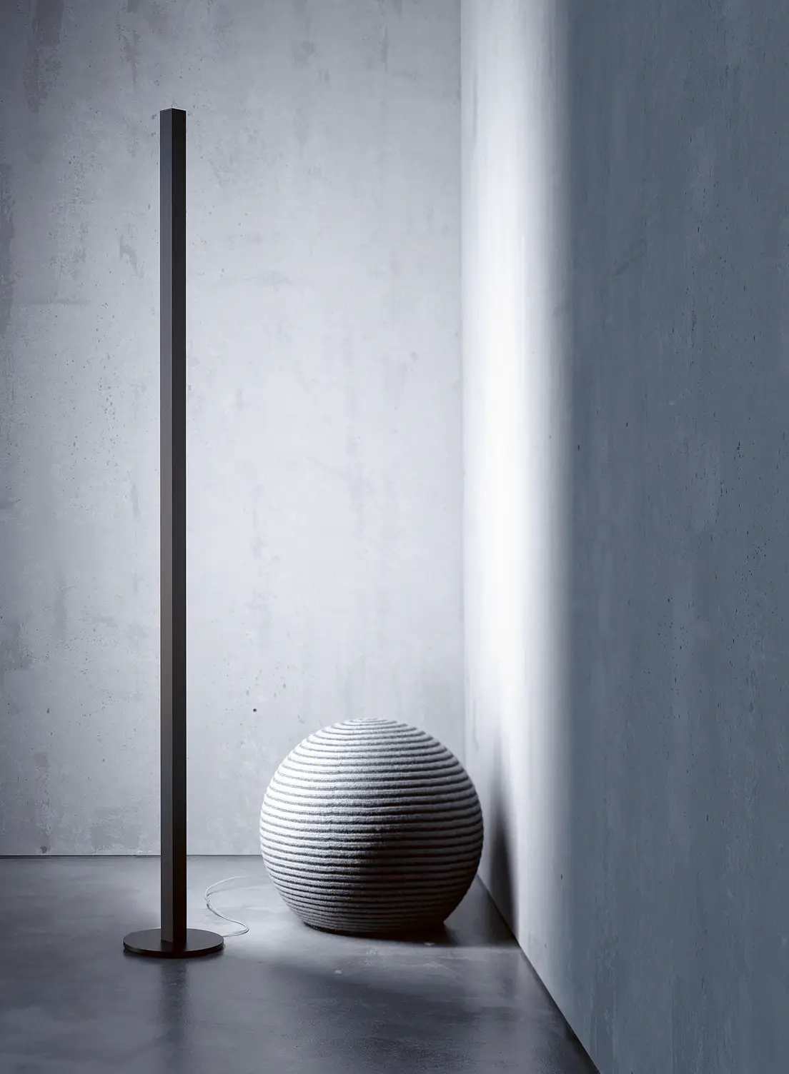 Schlanke schwarze Stehleuchte GERA Quadra Floor neben einem strukturierten, kugelförmigen Sitzmöbel in einer minimalistischen Raumecke.