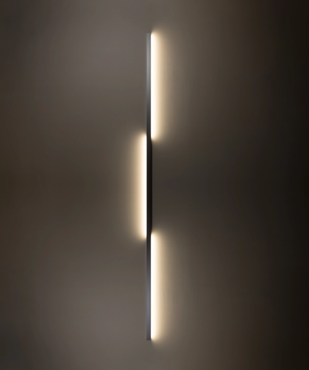 Eine minimalistische, vertikal angebrachte Wandleuchte, GERA Quadra Wall, dominiert das Bild vor einem dunklen Hintergrund. Ein schmale Aluminiumprofil erzeugt indirektes, warmes Licht, das rhythmisch versetzt austritt: oben und unten nach rechts, mittig nach links.